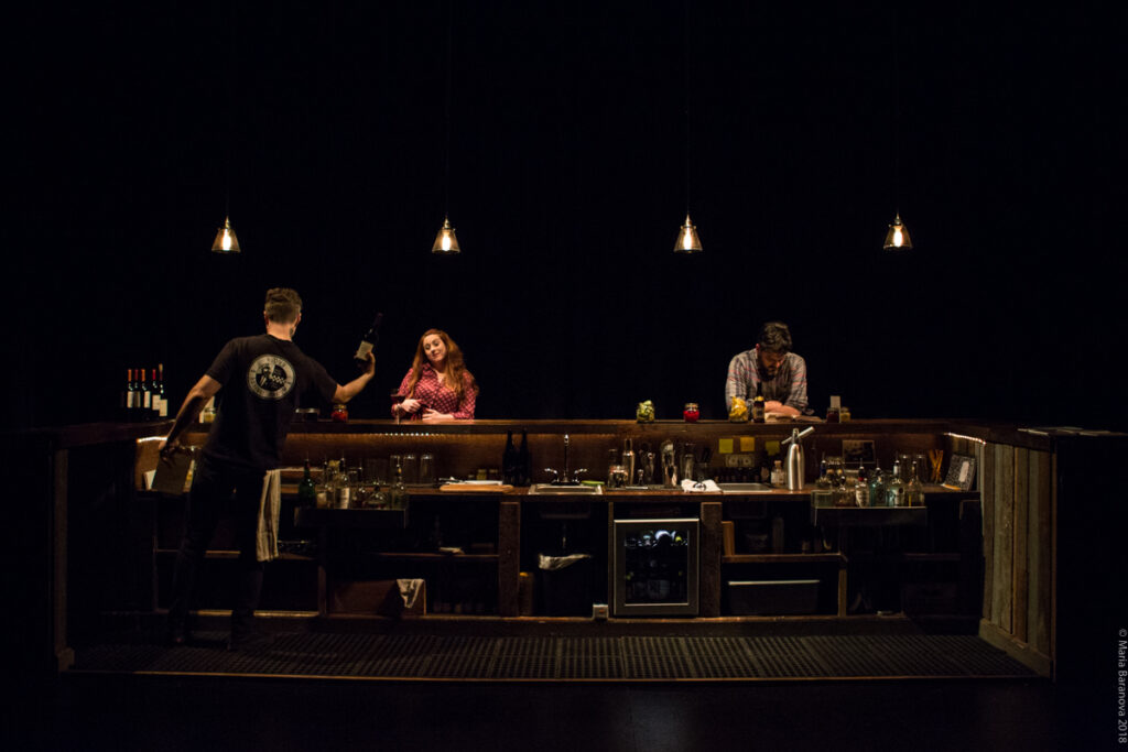 Photo of the play PORTO. A bartender is behind a bar, and a man and woman are sitting apart at the bar.